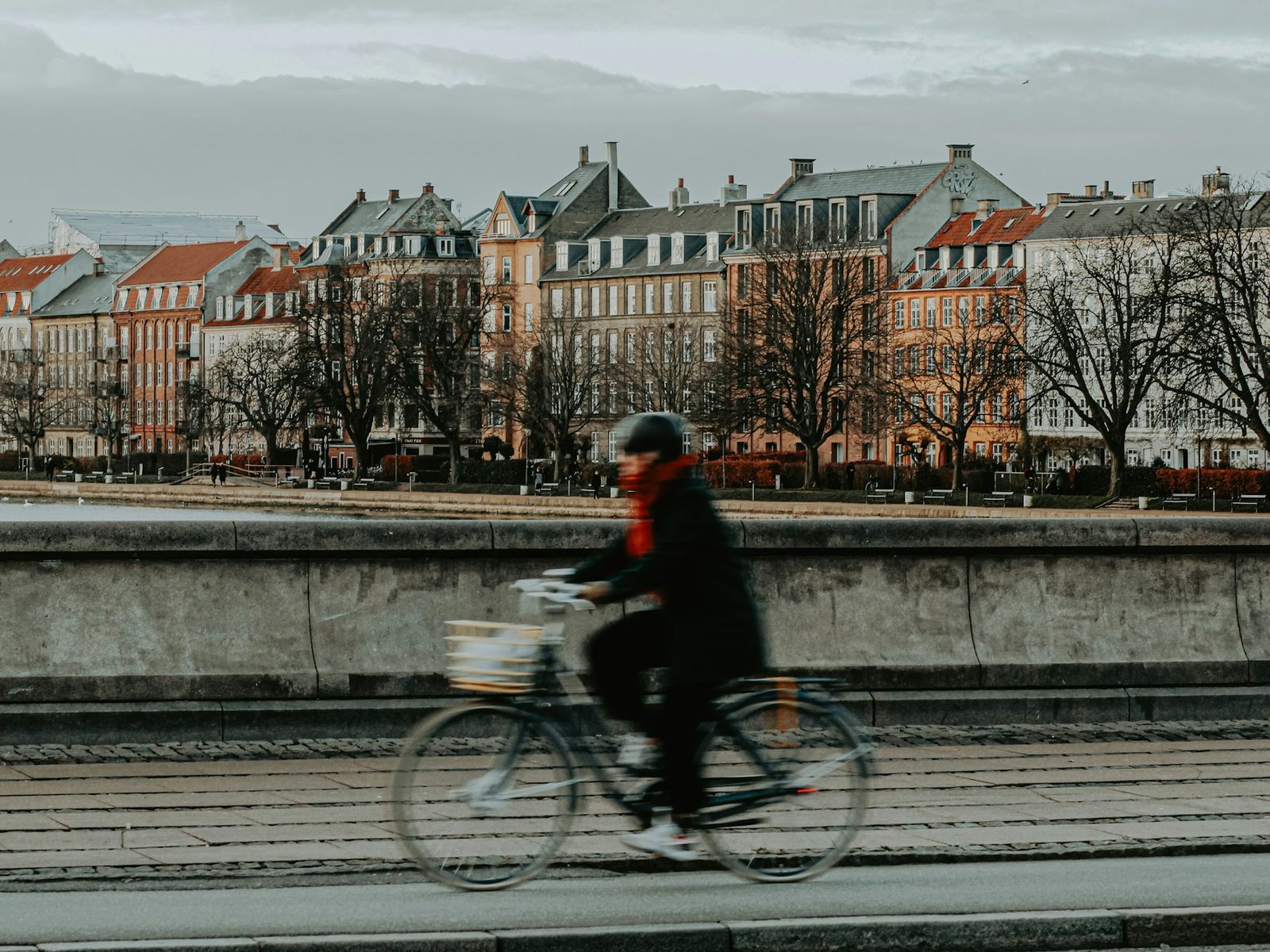 A cyclist in a red scarf rides past the bridge parapet over the Lakes, terraced houses and a grey sky behind.
